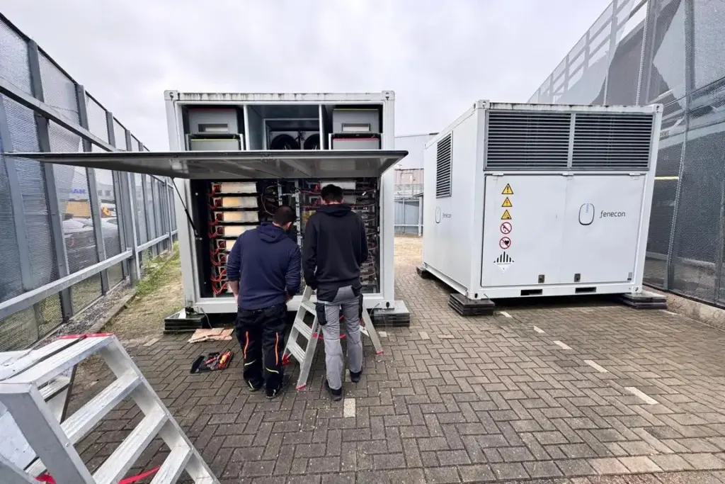 Zwei Techniker stehen vor einem geöffneten FENECON Industrial M Energiespeicher mit sichtbaren Batteriemodulen; daneben ein weiterer geschlossener Container mit FENECON-Logo und Warnsymbolen