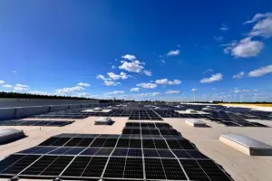 Großflächige Photovoltaikanlage auf dem Flachdach eines Industriegebäudes bei strahlend blauem Himmel, Windräder im Hintergrund sichtbar