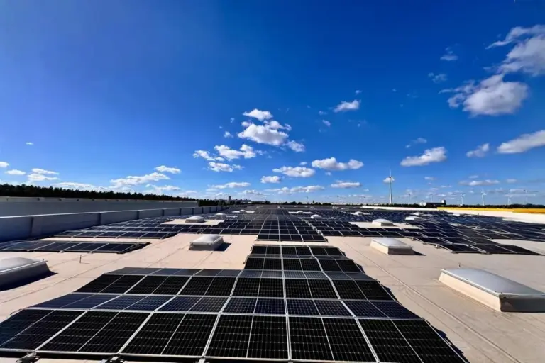 Großflächige Photovoltaikanlage auf dem Flachdach eines Industriegebäudes bei strahlend blauem Himmel, Windräder im Hintergrund sichtbar